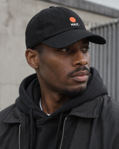 Man wearing the HALT. Dot Cap, a black low-profile baseball cap with coral dot logo, styled with a black hoodie